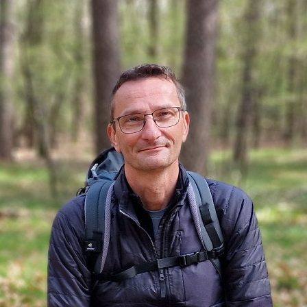 Mann mit Rucksack in einem Wald, trägt eine Brille und lächelt.