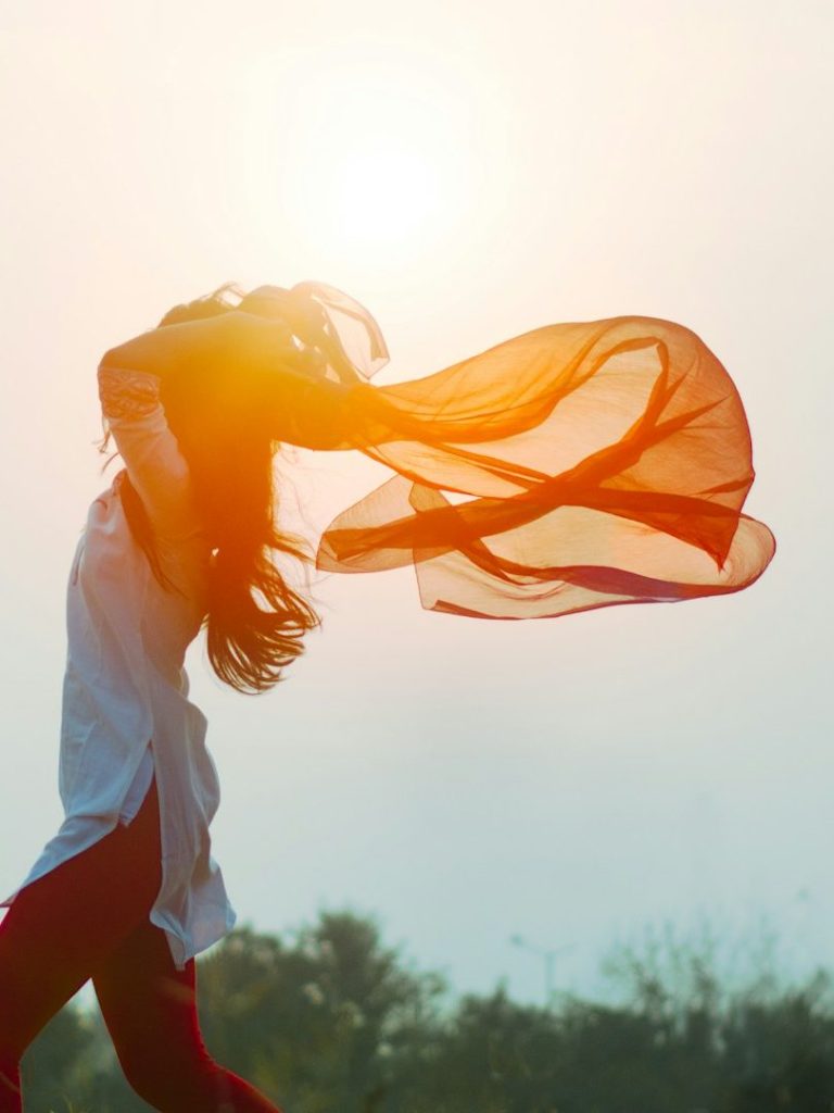 Junge Frau mit wehenden Haaren und einem roten Tuch vor der Sonne.