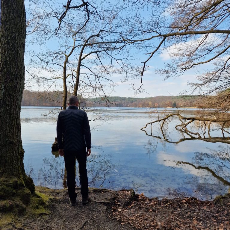 Eine Person steht am Ufer eines ruhigen Sees, umgeben von Bäumen und Wasser.